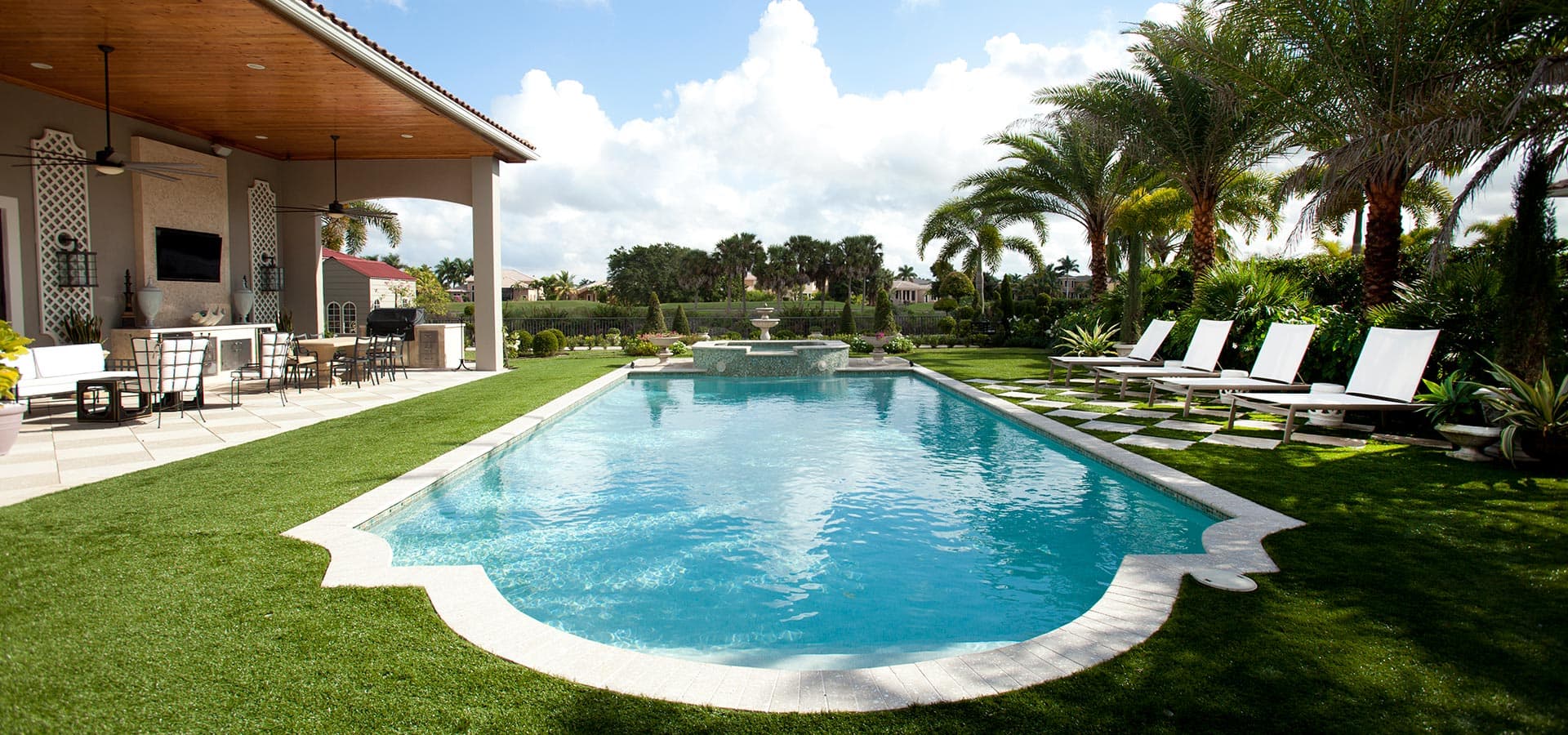 Pirela Atelier Interiors. Tropical backyard with a rectangular pool and raised spa fountain, a covered outdoor kitchen, and palm-lined chaise loungers. Boca Raton, Florida.