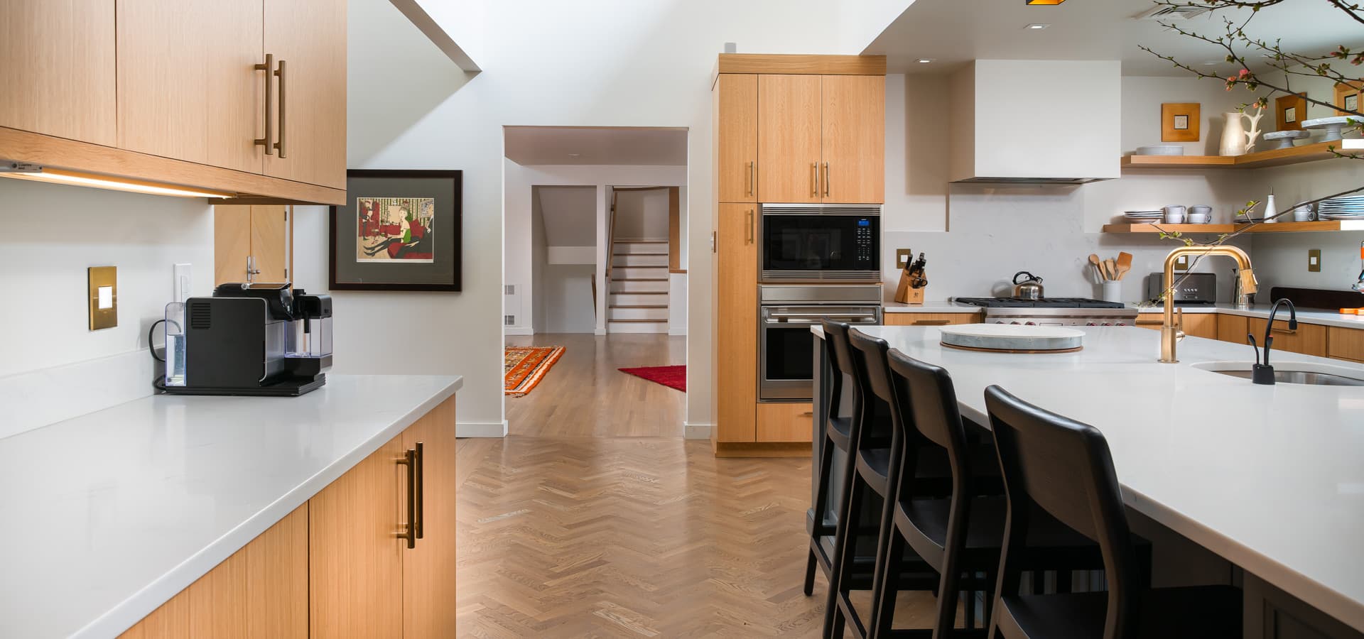 Pirela Atelier Interiors. Modern kitchen with light oak cabinetry, a white quartz island with black leather stools, and herringbone floors. Red Bank, New Jersey.